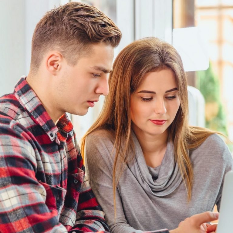 A young man and woman sit closely, looking at a smartphone together.